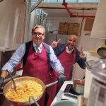 Due uomini in grembiule bordò preparano pasta al pomodoro in una cucina da campo improvvisata all'interno dello stand fieristico.