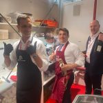 Tre uomini in abbigliamento da servizio posano dietro un bancone in cucina all'interno di uno stand fieristico con salumi e vino.