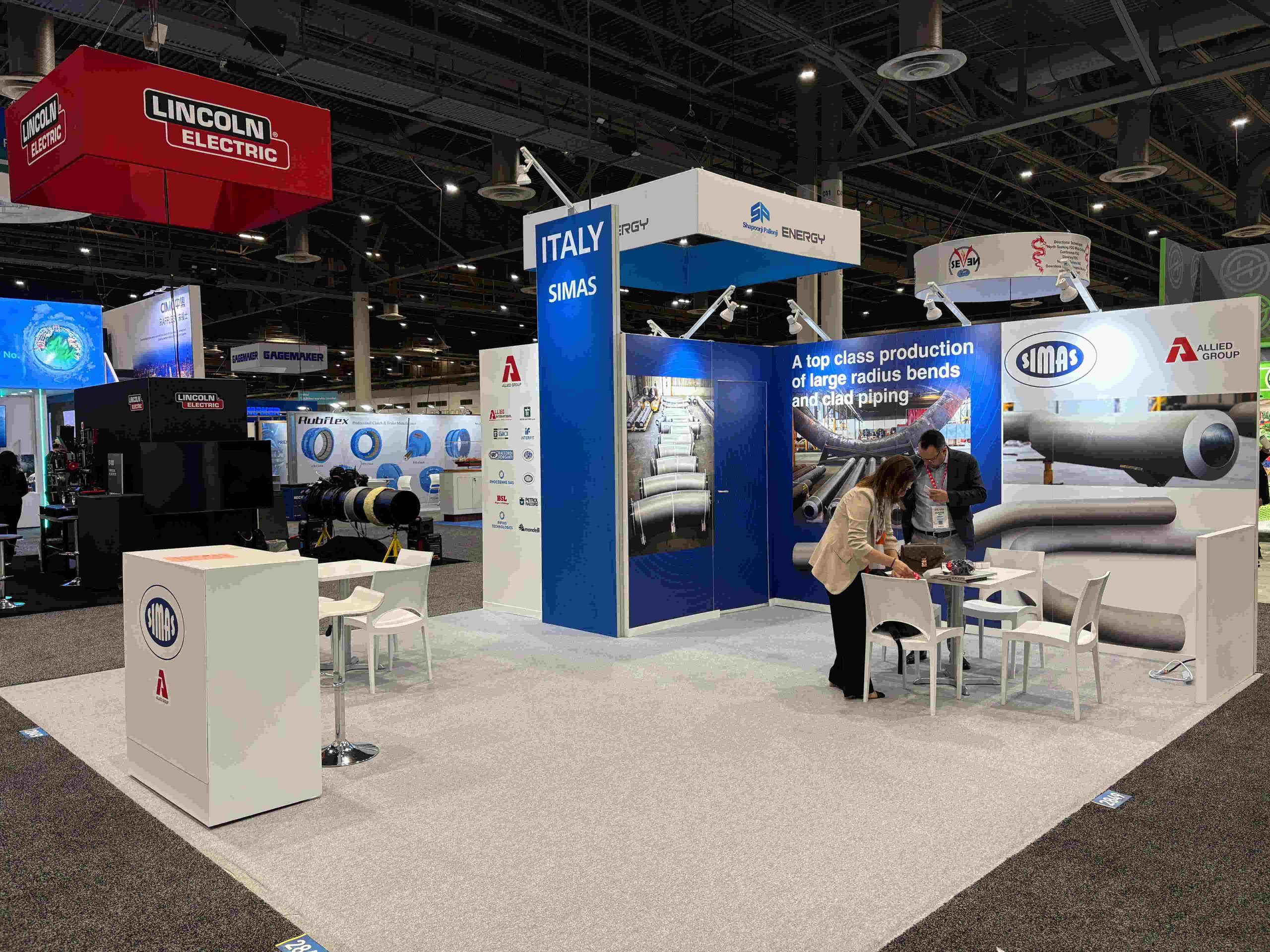 Stand fieristico SIMAS con pannelli informativi su tubazioni industriali durante l'evento OTC a Houston.